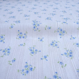 May include: White fabric with a ribbed texture and a pattern of small blue floral clusters with green leaves. The fabric has a soft, delicate appearance, suitable for various sewing projects.