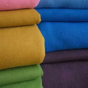 Puede incluir: Primer plano de tela doblada en varios colores. Los colores incluyen rosa, azul, dorado, verde y morado. La tela parece ser un material de punto, posiblemente algod&oacute;n o una mezcla de algod&oacute;n. Los pliegues crean un efecto de capas, mostrando los diferentes tonos.