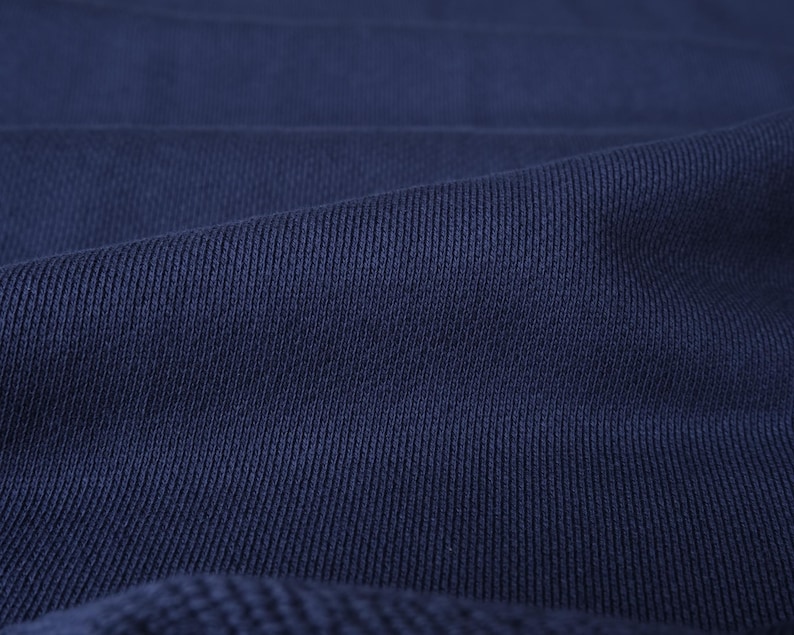 K&ouml;nnte beinhalten: Nahaufnahme eines marineblauen Rippstrickstoffs. Die strukturierte Oberfl&auml;che des Stoffes weist vertikale Linien und feine Falten auf, die ein Gef&uuml;hl von Tiefe erzeugen. Die tiefe, satte blaue Farbe eignet sich f&uuml;r Kleidung oder Heimtextilien.
