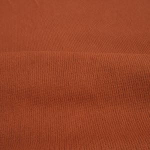 K&ouml;nnte beinhalten: Nahaufnahme eines strukturierten, rostfarbenen Stoffes. Das Material hat ein Rippenmuster mit vertikalen Linien &uuml;ber die Oberfl&auml;che. Der Stoff ist gefaltet, wodurch weiche Schatten entstehen und die Textur hervorgehoben wird.