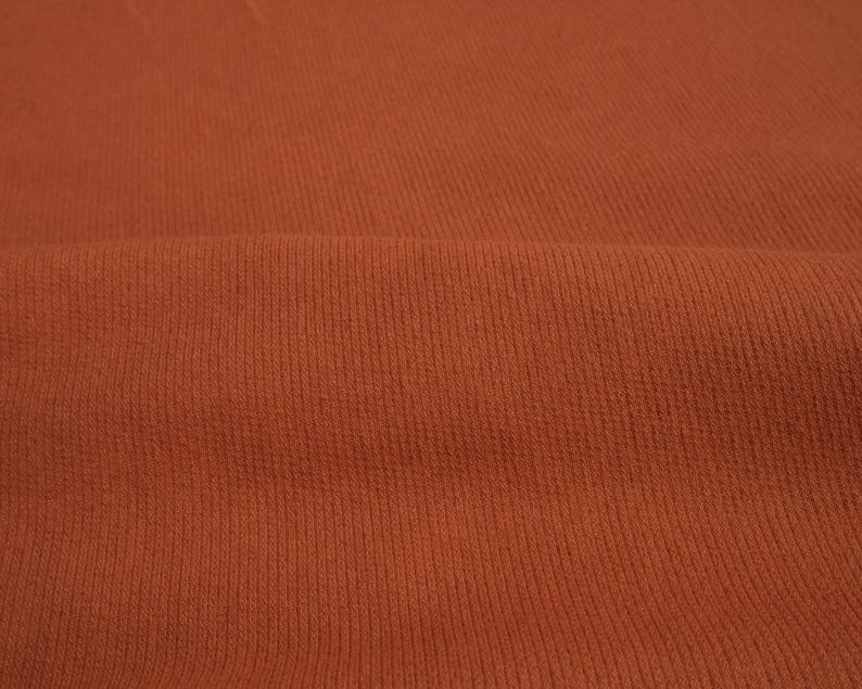 K&ouml;nnte beinhalten: Nahaufnahme eines strukturierten, rostfarbenen Stoffes. Das Material hat ein Rippenmuster mit vertikalen Linien &uuml;ber die Oberfl&auml;che. Der Stoff ist gefaltet, wodurch weiche Schatten entstehen und die Textur hervorgehoben wird.