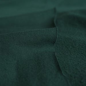 Pode incluir: Close-up de tecido verde escuro, mostrando duas texturas distintas. Um lado apresenta um tecido de malha liso e apertado, enquanto o outro mostra uma superf&iacute;cie texturizada e felpuda. O tecido est&aacute; dobrado.