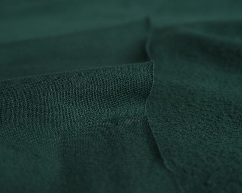 Pode incluir: Close-up de tecido verde escuro, mostrando duas texturas distintas. Um lado apresenta um tecido de malha liso e apertado, enquanto o outro mostra uma superf&iacute;cie texturizada e felpuda. O tecido est&aacute; dobrado.