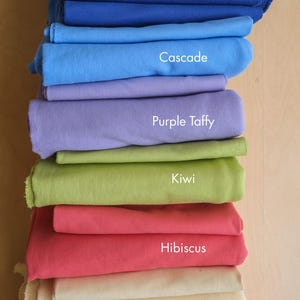 K&ouml;nnte beinhalten: Ein Stapel gefalteter Stoffe in verschiedenen Farben, darunter Deep Sea, Mushroom, Lapis, Cascade, Purple Taffy, Kiwi, Hibiskus, Butter, Persimmon und Bubblegum. Jeder Stoff ist mit seinem Farbnamen beschriftet.