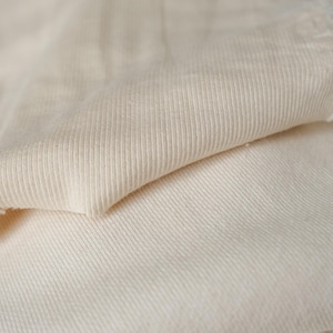 K&ouml;nnte beinhalten: Nahaufnahme eines cremefarbenen Rippstrickstoffs. Der Stoff ist gefaltet und zeigt die Textur und die rohe Kante des Materials. Der Strick hat vertikale Linien und der Stoff wirkt weich und leicht strukturiert.