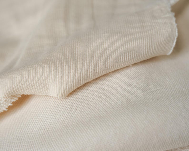 K&ouml;nnte beinhalten: Nahaufnahme eines cremefarbenen Rippstrickstoffs. Der Stoff ist gefaltet und zeigt die Textur und die rohe Kante des Materials. Der Strick hat vertikale Linien und der Stoff wirkt weich und leicht strukturiert.