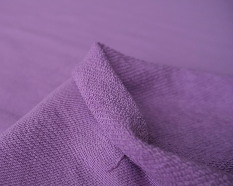 Puede incluir: Primer plano de un trozo de tela lavanda doblada. La tela tiene un aspecto texturizado y tejido, con un patr&oacute;n de costillas visible. Los bordes est&aacute;n doblados, revelando un borde m&aacute;s grueso y texturizado.