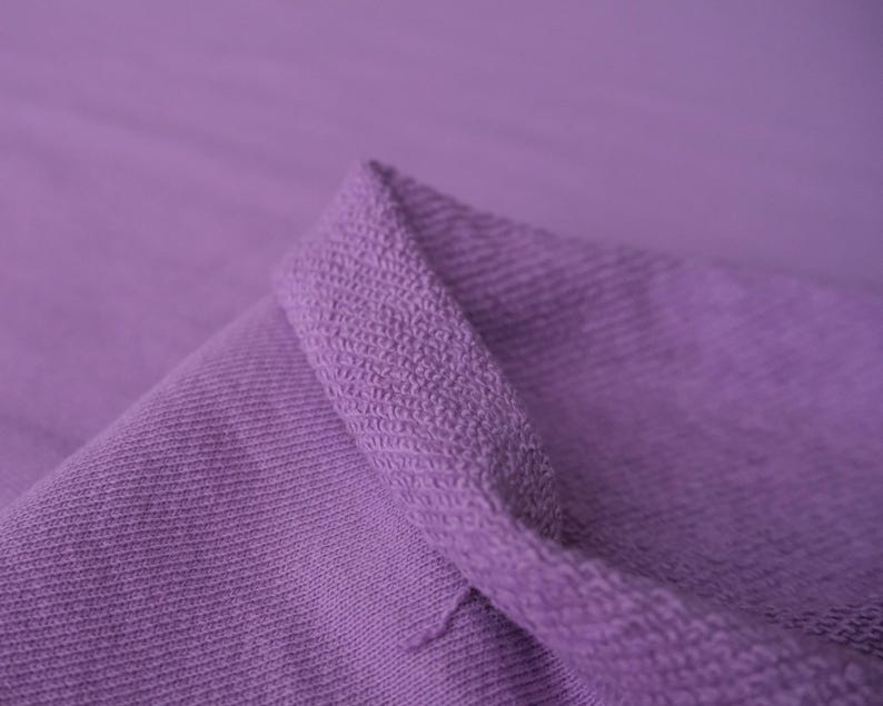 K&ouml;nnte beinhalten: Nahaufnahme eines gefalteten, lavendelfarbenen Stoffes. Der Stoff hat eine strukturierte, gestrickte Optik mit einem sichtbaren Rippenmuster. Die Kanten sind umgeschlagen und zeigen einen dickeren, strukturierteren Rand.