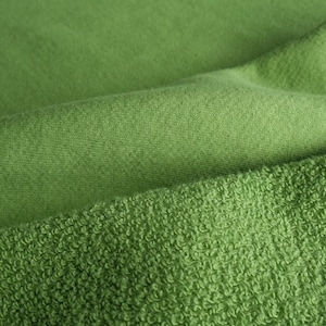 Puede incluir: Primer plano de tela verde con diferentes texturas. La capa superior es lisa, mientras que la capa inferior tiene una textura de rizo en bucle. La tela es de un color verde vibrante y s&oacute;lido, con pliegues y sombras suaves.