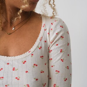 May include: A white long-sleeved shirt with a cherry print. The shirt has a scoop neck and a ribbed texture.