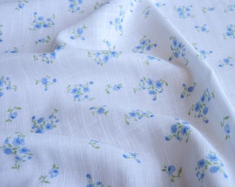 Op de afbeelding: Witte stof met een patroon van kleine blauwe bloemen en groene bladeren. De stof is licht gekreukt, wat de textuur benadrukt. Het bloemmotief is gelijkmatig verdeeld over het oppervlak.