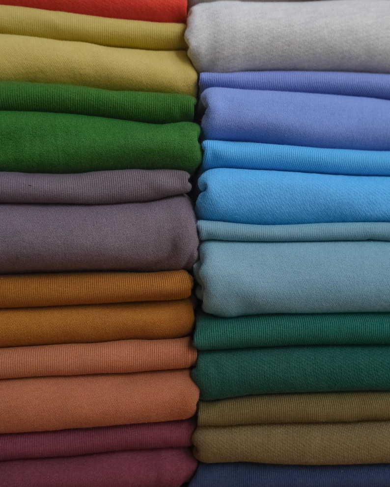 Pode incluir: Uma cole&ccedil;&atilde;o de tecidos dobrados em v&aacute;rias cores, incluindo vermelho, amarelo, verde, roxo, laranja e azul. O tecido parece ser um material de malha macia, cuidadosamente empilhado em fileiras. A imagem mostra um espectro de tons, de quentes a frios.