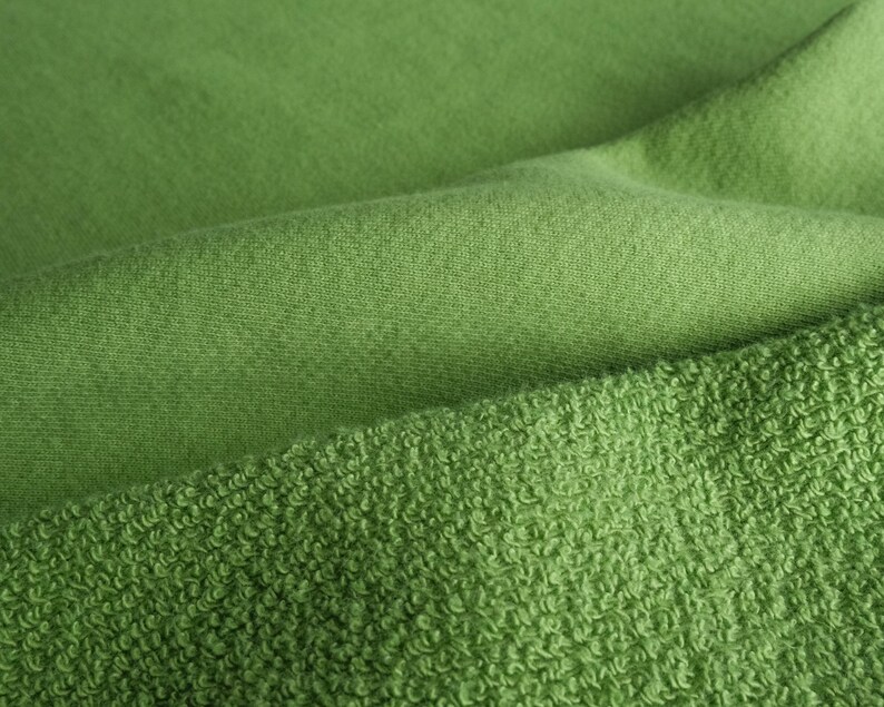 K&ouml;nnte beinhalten: Nahaufnahme von gr&uuml;nem Stoff mit unterschiedlichen Texturen. Die obere Schicht ist glatt, w&auml;hrend die untere Schicht eine geschlungene, frotteeartige Textur aufweist. Der Stoff ist ein leuchtender Gr&uuml;nton mit weichen Falten und Schatten.