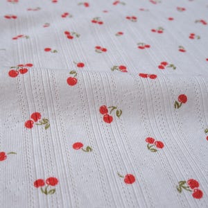 May include: White fabric with a repeating pattern of red cherries. The fabric has a textured, ribbed surface.