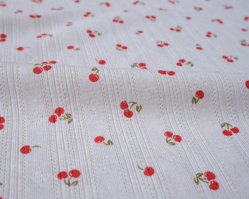 May include: White fabric with a repeating pattern of red cherries. The fabric has a textured, ribbed surface.