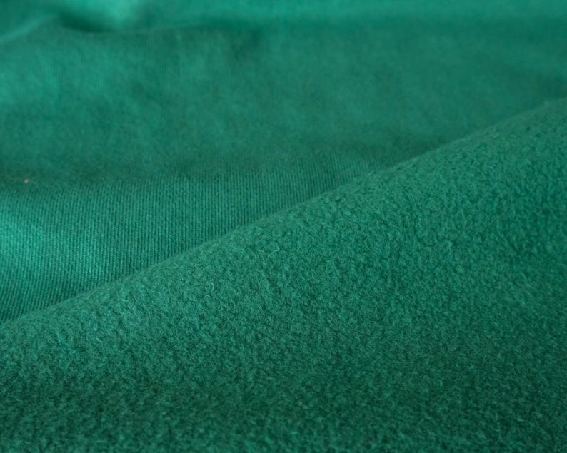 Pode incluir: Close-up de um tecido verde esmeralda texturizado. O tecido tem uma textura macia e ligeiramente difusa de um lado e uma textura canelada do outro. O tecido est&aacute; dobrado, criando sombras e profundidade.