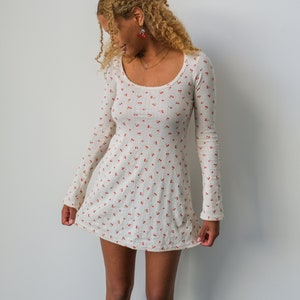 May include: A white mini dress with a cherry print and long sleeves. The dress has a scoop neckline and a fitted silhouette.