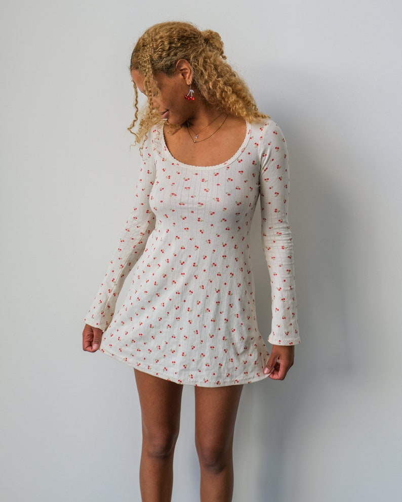 May include: A white mini dress with a cherry print and long sleeves. The dress has a scoop neckline and a fitted silhouette.