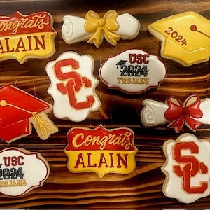 Może przedstawiać: Zestaw dziewięciu ozdobnych ciasteczek cukrowych z motywami związanymi z ukończeniem studiów. Ciasteczka są ozdobione lukrem w kolorze czerwonym, żółtym i białym z napisem "Congrats Alain", "USC 2024 Trojans", "2024" i "SC".