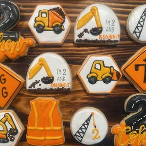 Può includere: Una collezione di biscotti di zucchero decorati a tema costruzione. I biscotti presentano immagini di veicoli da costruzione, segnali stradali e il numero "2". Alcuni biscotti hanno il testo "I'm 2 and digging it!" e il nome "Cohen".