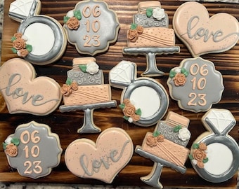 Puede incluir: Una colección de 12 galletas de azúcar decoradas con varios diseños, incluyendo corazones, anillos, pasteles y fechas. Las galletas están decoradas con glaseado rosa, gris y plateado y presentan las palabras "love" y "06 10 23".