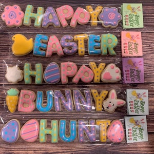 Puede incluir: Un conjunto de cinco galletas de azúcar decoradas que deletrean "Happy Easter Bunny Hunt". Cada galleta está decorada con diferentes colores pastel y patrones.