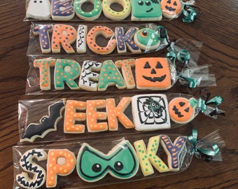 May include: Five clear plastic bags each containing a set of Halloween-themed sugar cookies. The cookies are decorated with various colors and designs, including ghosts, bats, pumpkins, and spiders. The bags are tied with green and white ribbon.