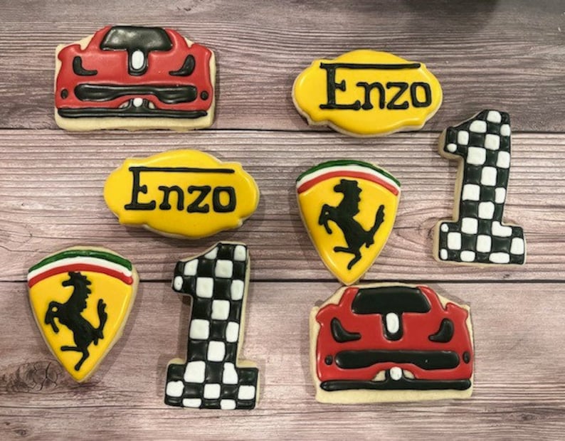 May include: A set of nine decorated sugar cookies featuring a red and black race car, a yellow and black checkered flag, a yellow and black checkered number one, a yellow and black name tag with the name Enzo, a red and black Ferrari logo, and a red and black race car.