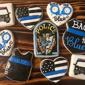 Könnte beinhalten: Sortiment von dekorierten Zuckerkeksen mit einem blauen Linien-Motiv. Die Kekse zeigen Polizeiautos, Handschellen, Herzen und ein Polizeibadge. Die Kekse sind mit schwarzem, weißem und blauem Zuckerguss verziert.