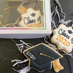 Puede incluir: Una caja blanca con una ventana transparente que muestra dos galletas con forma de gorro de graduación. Una galleta es negra con texto blanco que dice "2020". La otra galleta es blanca con texto negro que dice "Class of 2020". La caja está sobre una superficie negra con papel triturado blanco y negro.