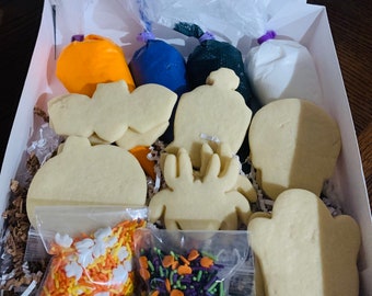 May include: A white box filled with Halloween cookie decorating supplies. The box contains six sugar cookies in various shapes, including a pumpkin, a spider, a ghost, and a bat. There are also three bags of icing in blue, green, and white, and two bags of sprinkles in orange and green.