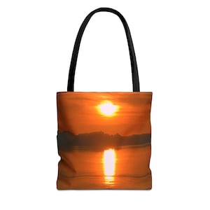 May include: A tote bag featuring a vibrant sunset scene with a bright orange sky, a glowing sun, and a dark silhouette of a distant landscape reflected on the water surface. The bag has two solid black shoulder straps attached to the top edge.
