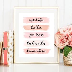 May include: A framed print with a white background and pink watercolor brushstrokes. The text on the print reads "risk-taker, hustler, girl-boss, hard-worker, dream-chaser".