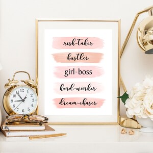 May include: A framed print with a white background and pink watercolor stripes. The text on the print reads "risk-taker", "hustler", "girl-boss", "hard-worker", and "dream-chaser".