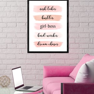 May include: A black framed print with pink and peach watercolor brushstrokes behind the words "risk-taker", "hustler", "girl-boss", "hard-worker", and "dream-chaser". The print is hanging on a white brick wall above a pink sofa.