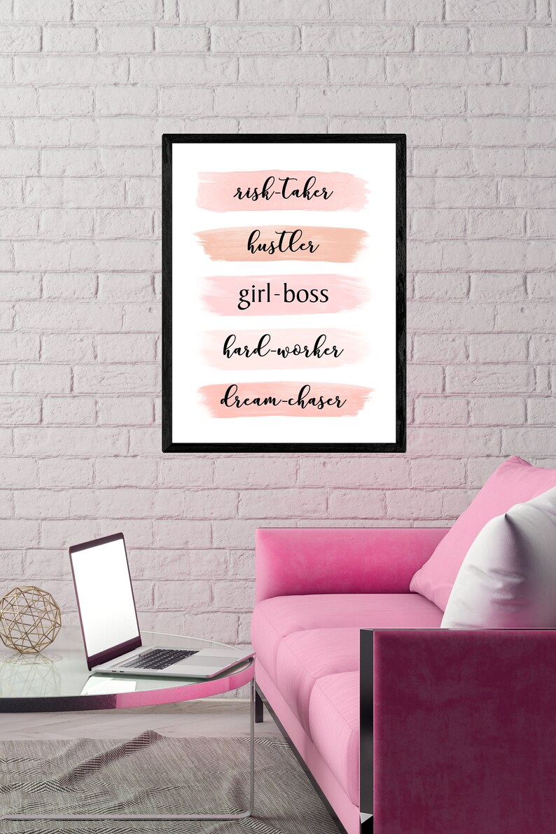 May include: A black framed print with pink and peach watercolor brushstrokes behind the words "risk-taker", "hustler", "girl-boss", "hard-worker", and "dream-chaser". The print is hanging on a white brick wall above a pink sofa.