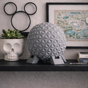 Puede incluir: Una escultura esférica geométrica gris con un patrón triangular, apoyada en una estantería. Junto a ella hay una maceta con forma de calavera con una suculenta, una silueta de Mickey Mouse y un mapa enmarcado. También hay un bolígrafo y libros en la estantería.