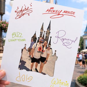 Puede incluir: Un recorte de papel blanco de un castillo con una foto de una pareja en su interior. El papel tiene firmas de personajes de Disney en tinta roja, morada, amarilla y verde, incluyendo "Ariel", "Mickey Mouse" y "Buzz Lightyear".