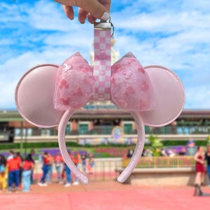 May include: Pink mouse ear headband with matching bow and a checkered lanyard. The ears are round and made of a solid pink material. The bow is made of a sheer pink fabric with small mouse silhouettes. The lanyard is pink and white checkered.