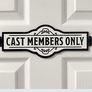 Könnte beinhalten: Ein rechteckiges Schild mit dem Text "CAST MEMBERS ONLY" in fetten, schwarzen Buchstaben. Das Schild hat einen weißen Hintergrund mit einem dekorativen schwarzen Rand und Verzierungen. Es ist an einer weißen Tür angebracht.