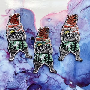 May include: Three embroidered iron-on patches featuring a brown bear with a mountain scene inside. The bear is standing on its hind legs with its mouth open. The mountain scene includes a sunset, trees, and a river.