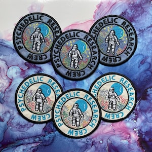 May include: Six embroidered patches with a psychedelic space theme. Each patch features a cartoon astronaut in a white spacesuit standing in front of a colorful nebula with a crescent moon. The text "Psychedelic Research Crew" is written in a circular pattern around the astronaut.