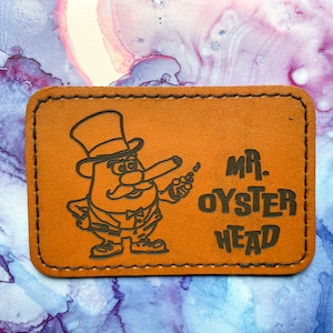 May include: A brown leather patch with a cartoon character wearing a top hat and holding a cigar. The text "MR. OYSTER HEAD" is printed in black.