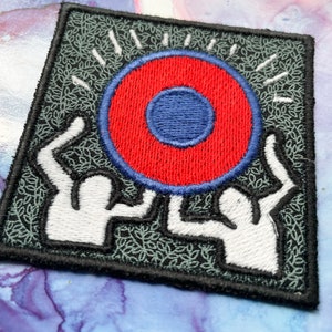 May include: A black and green embroidered patch with two white figures holding up a red circle with a blue center.