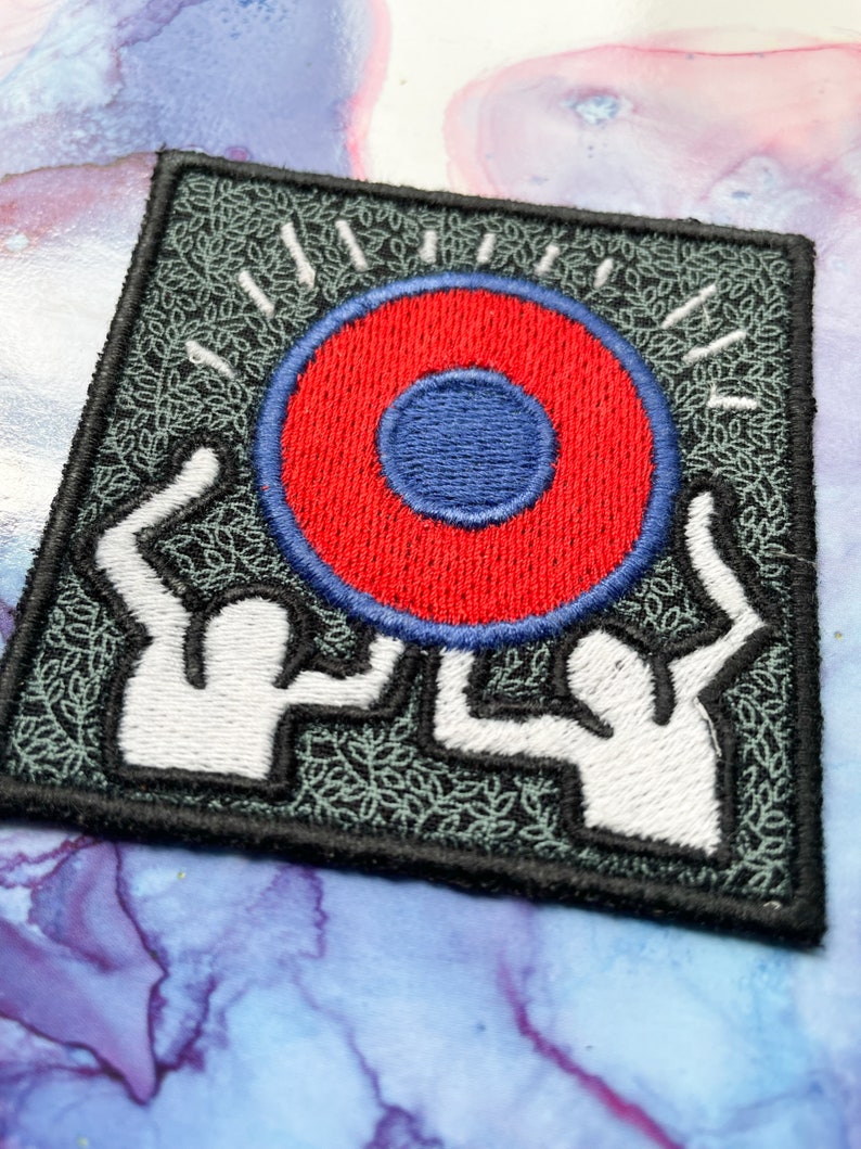 May include: A black and green embroidered patch with two white figures holding up a red circle with a blue center.