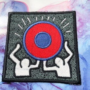May include: A black and white embroidered patch with two figures holding up a red and blue circle with a white outline. The background is a dark gray with a pattern of small white leaves.