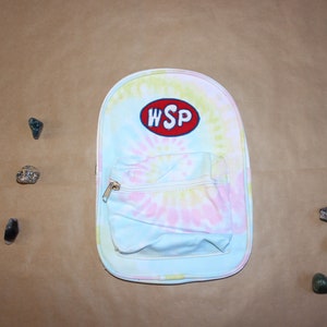 May include: A blue tie-dye backpack with a red oval patch that says "WSP" on it. The backpack has a small front pocket with a gold zipper.