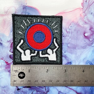 May include: Embroidered iron-on patch featuring two figures holding up a red and blue circle with a white outline. The background is gray with a black border.