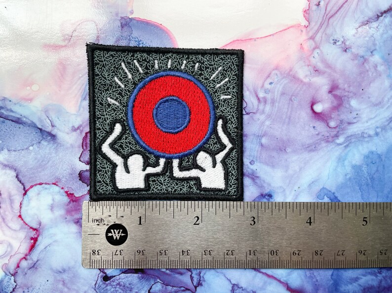 May include: Embroidered iron-on patch featuring two figures holding up a red and blue circle with a white outline. The background is gray with a black border.