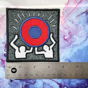 May include: A black and white embroidered patch featuring two figures holding up a red and blue circle with a smaller blue circle in the center. The circle has a white outline and is surrounded by white rays.